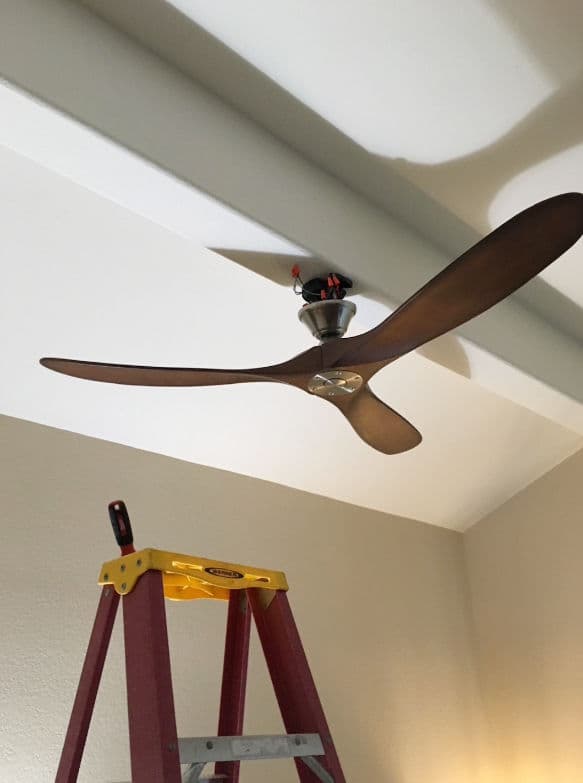 Ceiling fan installation in a room with a yellow ladder and wooden blades and a light fixture.