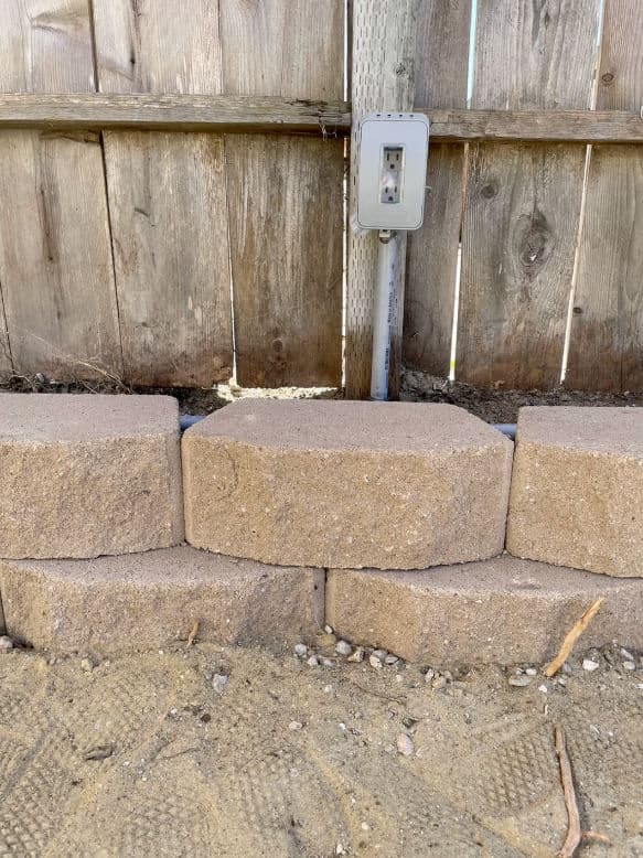 Retaining wall made of beige concrete blocks next to a wooden fence and electrical outlet.