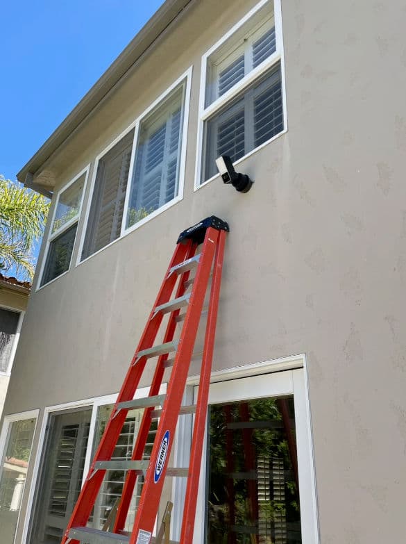 Red ladder leaning against a house with large windows and an outdoor security camera installed.